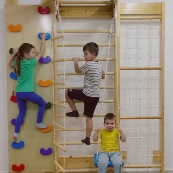 Trois enfants utilisant un kit espalier junior multifonction en bois : une fillette sur le mur d'escalade, un garçon grimpant sur l'espalier central avec échelle de corde, et un jeune garçon assis sur la balançoire intégrée